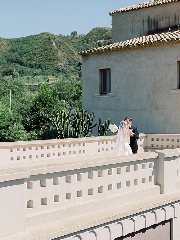 Wedding kiss as bride in a strapless gown holds a white bouquet, long veil flowing on a terrace balcony with cactus and hills beyond