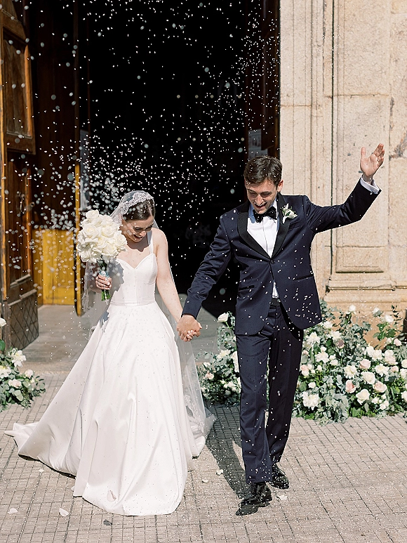 Wedding recessional as bride and groom exit the church doorway hand in hand, veil and bouquet lifted, rose petals tossed on stone steps