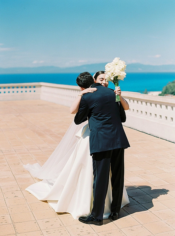 Wedding couple portrait of bride and groom hugging, bride holding a white bouquet on a seaside terrace with blue ocean and sky backdrop