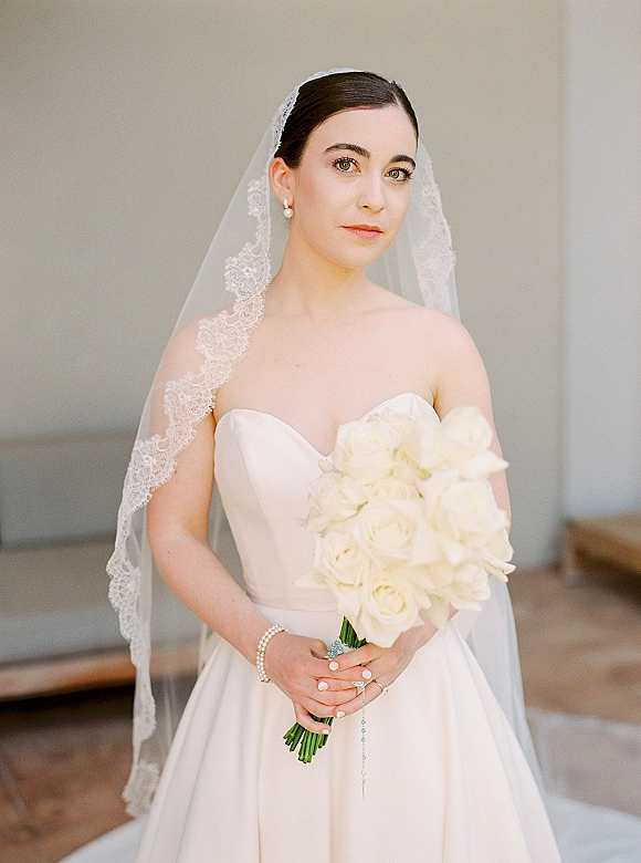 Bridal portrait of a bride holding bouquet of white roses, wearing a strapless dress and lace veil, seated by a wooden bench indoors