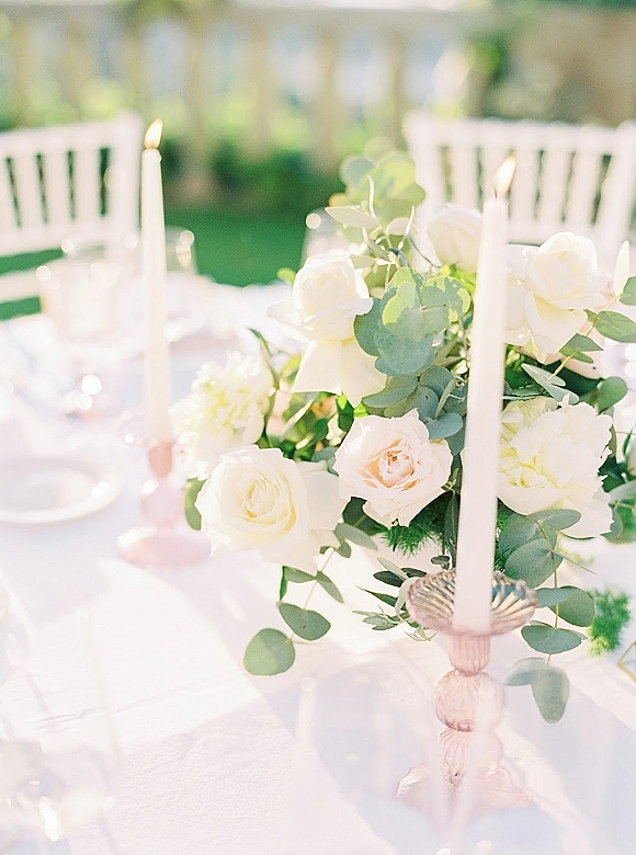 Reception tablescape with wedding table centerpiece of white roses and eucalyptus, taper candles and glassware on white linens in a garden setting