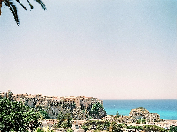 Coastal town view of a cliffside village with palm fronds and tiered buildings above turquoise sea, rocky shoreline, and open sky