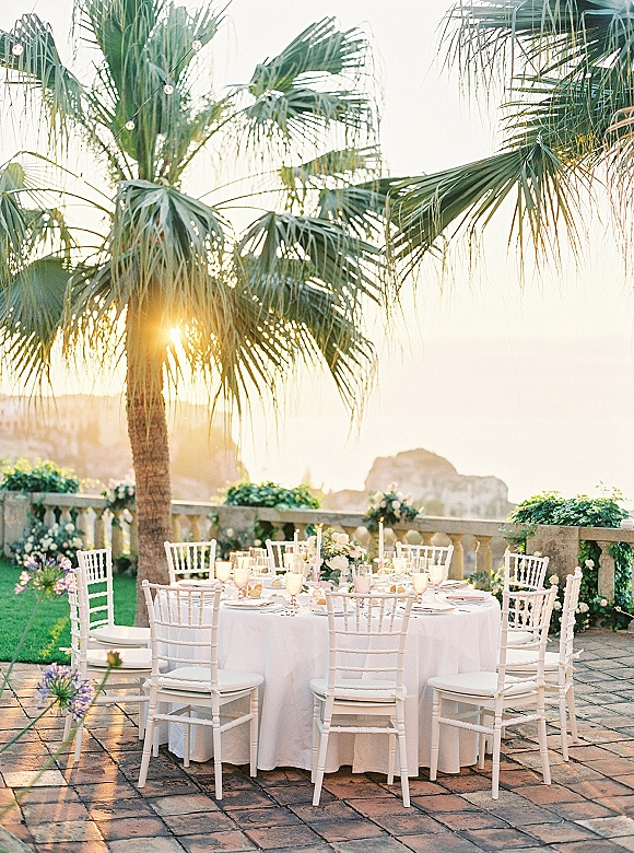 Reception tablescape with an outdoor reception table on a stone terrace, white linens, floral centerpiece, taper candles, and ocean sunset backdrop