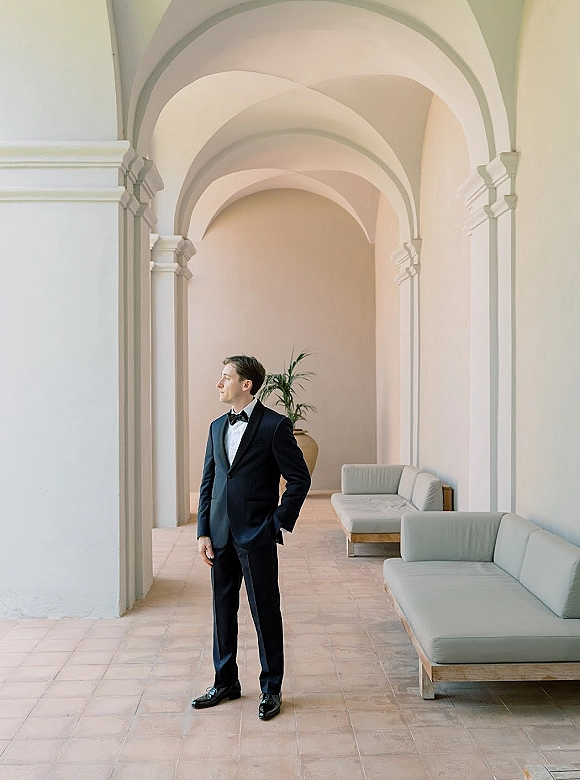 Groom portrait in a black tuxedo with bow tie and boutonniere, hands in pockets, standing in an arched hallway with columns