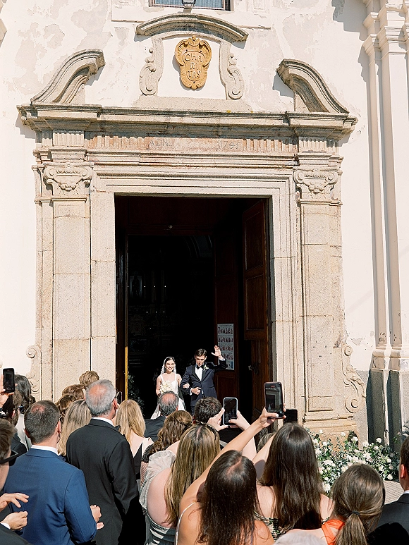 Wedding recessional as newlyweds exit church, bride in veil with bouquet beside groom in tuxedo, guests cheering with phones in sunlight