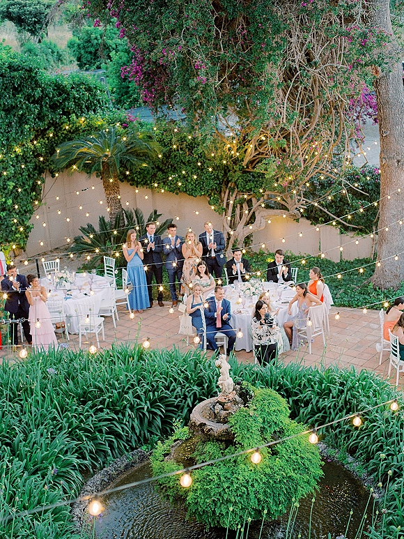 Wedding reception under string lights with round tables and white linens set on a brick patio in a lush garden courtyard by a fountain