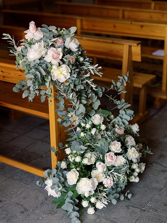 Ceremony aisle flowers with blush roses, peonies, and eucalyptus garland along wooden pews on a stone tile floor indoors