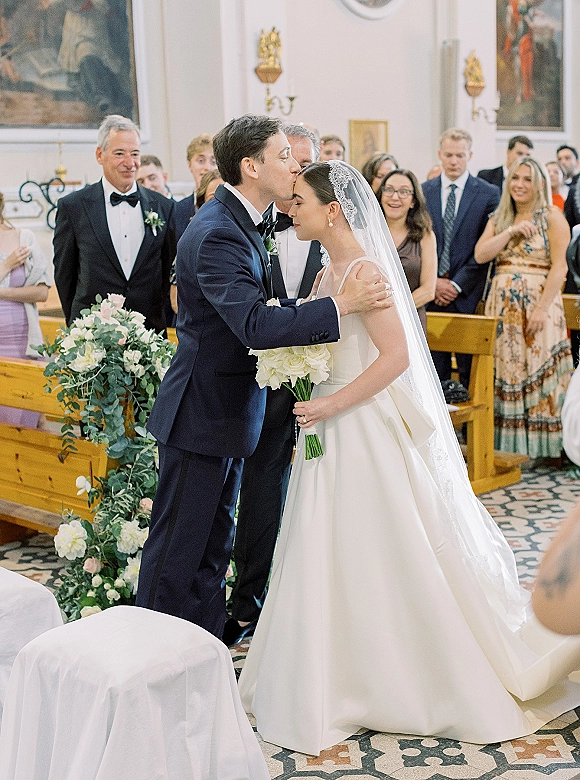 Ceremony moment as bride and groom embrace, groom kissing her forehead beside greenery-lined aisle in a classic church interior