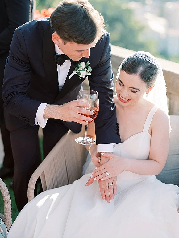 Couple portrait of bride and groom laughing as he leans over her, veil and tuxedo in sunlight on a green outdoor terrace