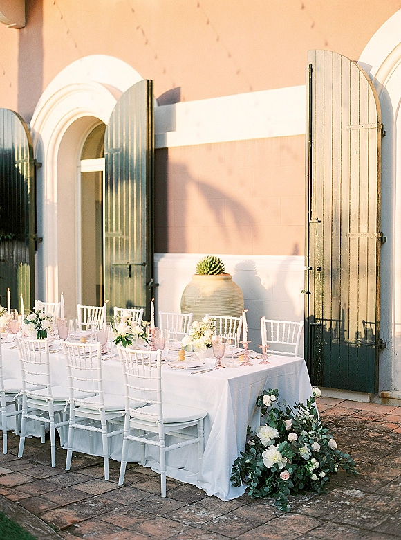 Reception tablescape with outdoor wedding tablescape details, white linens, pink goblets, taper candles and florals on a sunlit patio by green doors