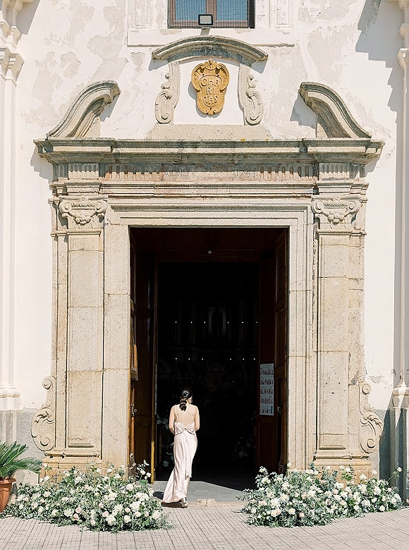 Bridal portrait of a bride walking into church in a satin bow-back dress, seen from behind at an ornate stone doorway with florals