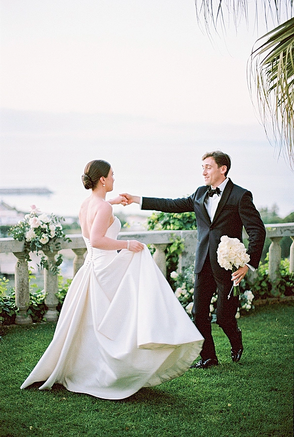 Couple portrait of bride in a strapless satin wedding dress and groom in tuxedo holding a white rose bouquet on an oceanfront terrace.