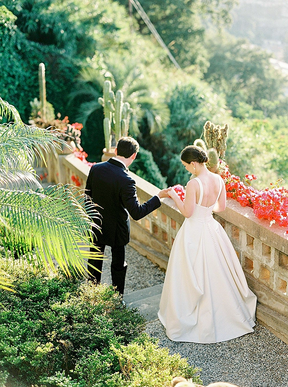 Couple portrait of bride and groom walking hand in hand down sunlit stone steps, her ballgown and his tuxedo framed by cactus garden greenery