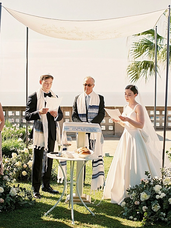 Wedding ceremony under a chuppah with floral arrangements as bride and groom read vows, oceanfront lawn with palm trees behind