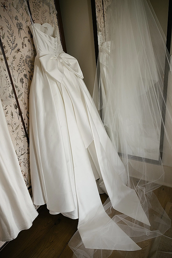 Wedding dress, a strapless satin gown with oversized bow and button back hanging with long veil beside a floor mirror on floral wallpaper