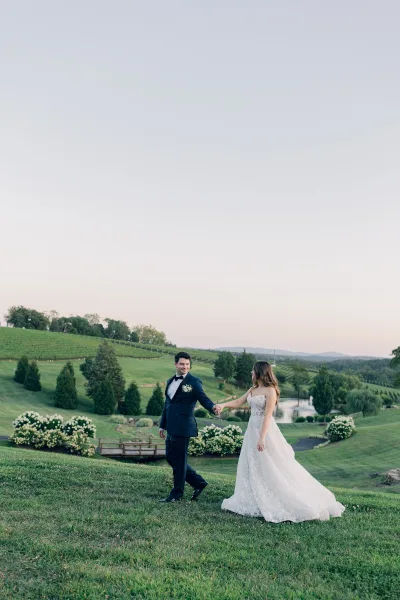 Couple portrait of the bride and groom walking along vineyard rows by a pond, her lace bridal gown flowing against rolling hills.