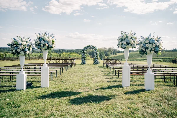 Outdoor ceremony setup for a vineyard wedding ceremony with a floral arch, facing vineyard rows and rolling hills beneath a blue sky.