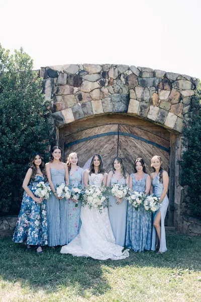 Bride and bridesmaids in blue bridesmaid dresses stand beneath a rustic stone arch with wooden doors, holding white and green bouquets.