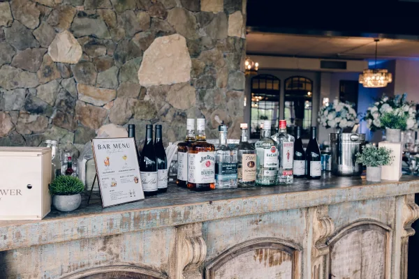 Wedding bar setup at a cocktail hour bar featuring a bar menu sign, set against a stone wall with chandeliers and mirrors.