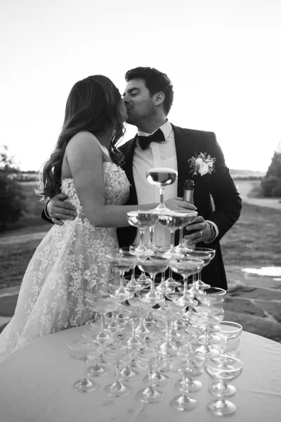 Wedding kiss portrait of the bride and groom kissing beside a gleaming champagne tower in an outdoor setting with trees beyond.