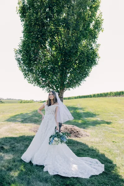 Bridal portrait of a bride with bouquet, standing beneath a tree by vineyard rows across an open field under the sky.