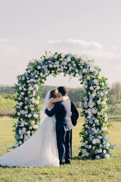 Wedding first kiss for a couple under a floral arch, on a sunlit lawn with open field and trees surrounding, capturing a tender ceremony moment.