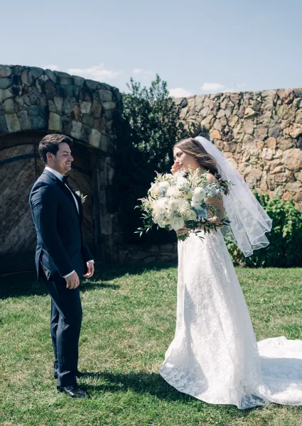 First look moment in a couple portrait as the bride in a lace gown approaches the groom by a stone wall and wooden door.