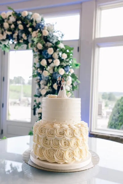 Wedding cake, a two tier wedding cake adorned with buttercream rosettes, set beneath a floral arch and large windows with a garden view.