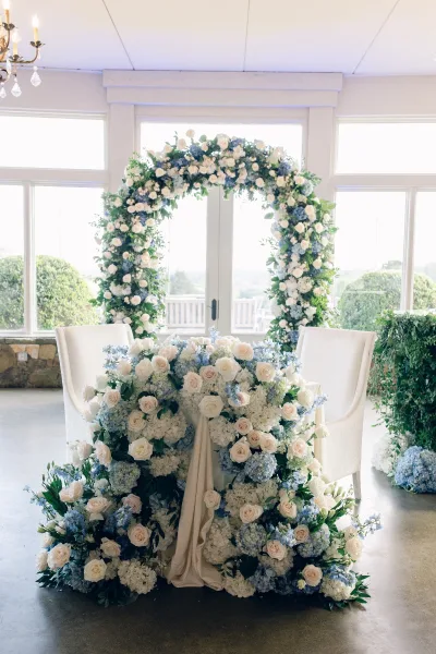 Sweetheart table decor framed by a floral arch backdrop, set before large windows overlooking garden shrubs and a stone wall.