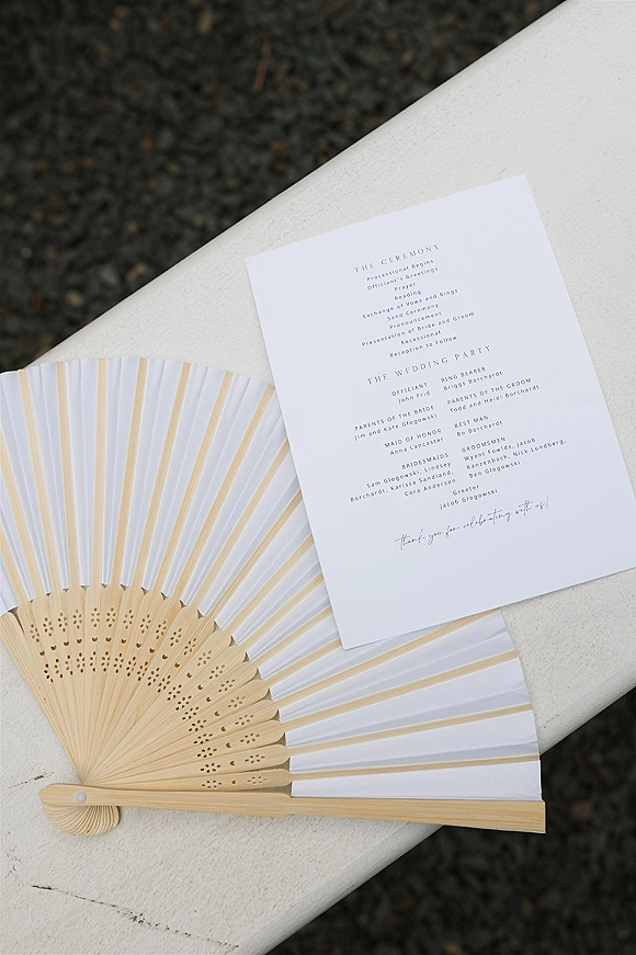 Wedding program card with minimal modern typography, attached to a paper fan, resting on a white bench over gravel ground