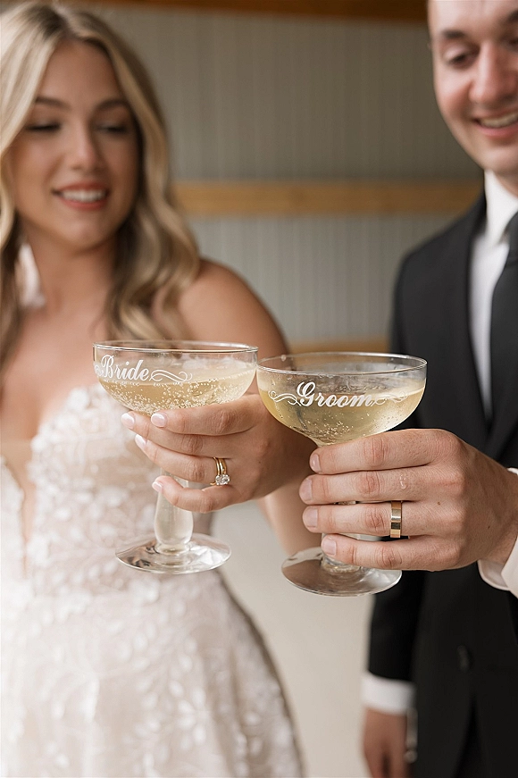 Wedding toast as bride and groom clink etched champagne coupe glasses, rings visible, against indoor wall paneling backdrop