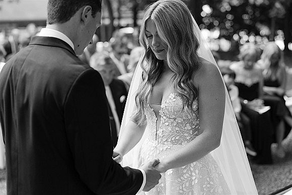 Ceremony moment at an outdoor wedding ceremony as bride in strapless lace dress and long veil holds groom’s hand before guests and trees