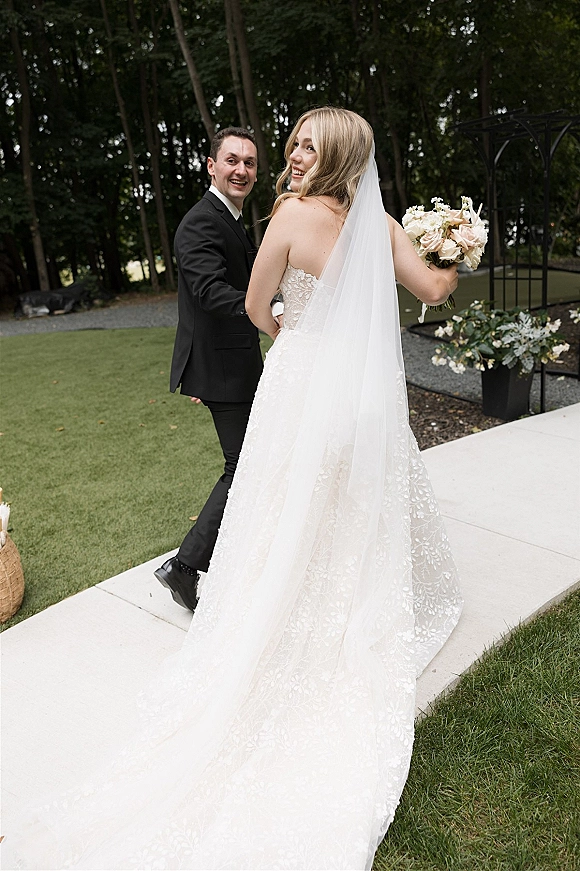 Couple portrait of bride looking back as newlyweds hold hands on a garden walkway, her cathedral veil flowing past a metal arch