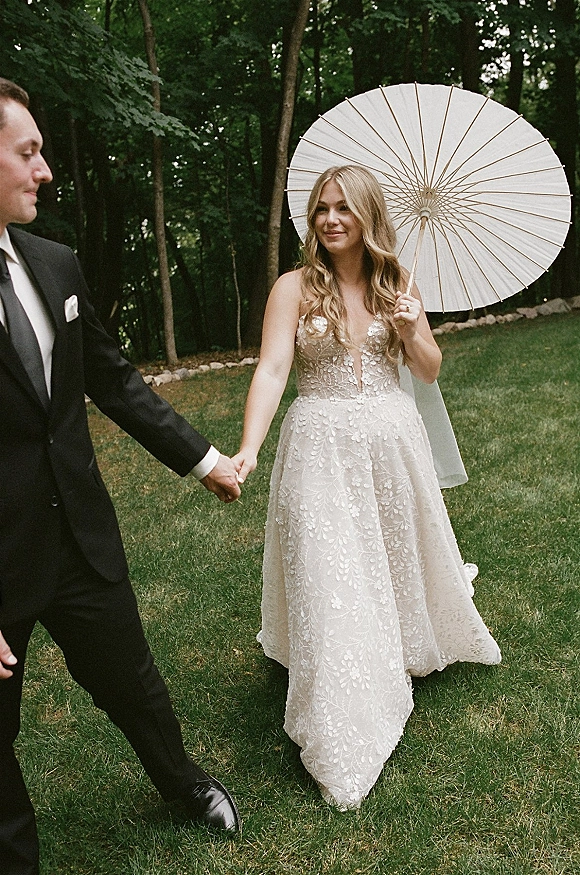 Couple portrait of bride and groom holding hands, bride in lace dress with veil and white parasol on a lawn by forest trees