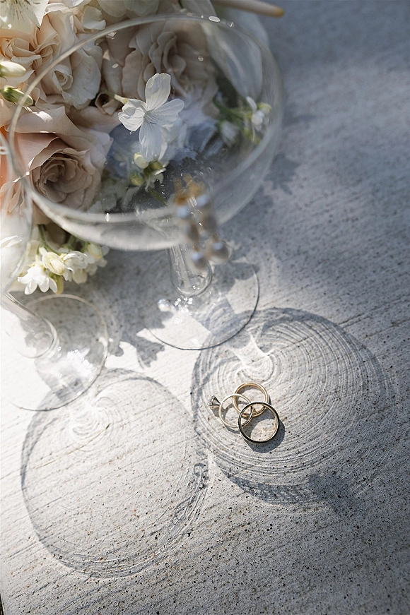 Wedding rings with a diamond engagement ring and band set on stone beside a champagne coupe, white blossoms, and soft natural light shadows