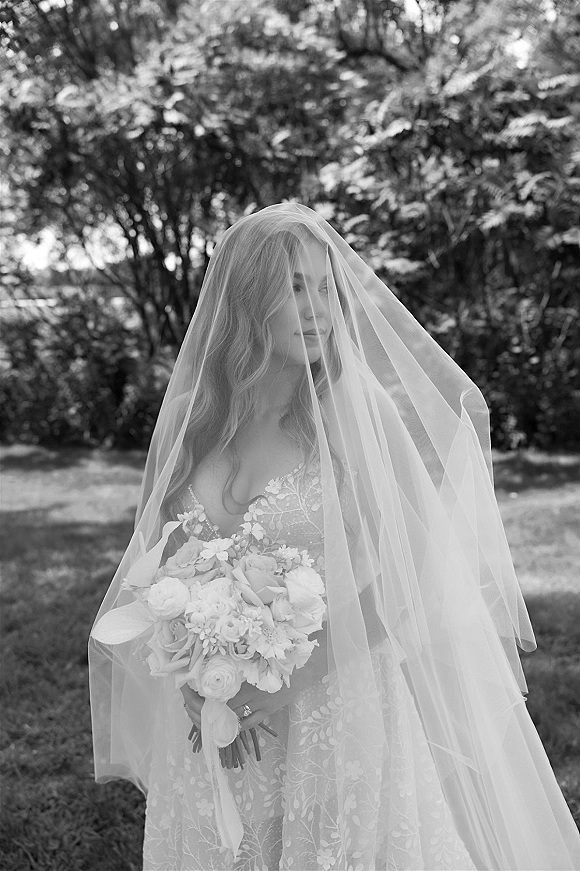 Bridal portrait in black and white of a bride with veil over her face holding a bouquet on a garden lawn with trees behind