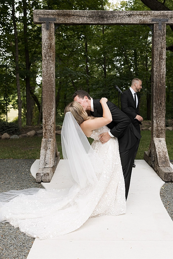 Wedding kiss at a rustic wedding arbor as the groom dips the bride in a lace dress, her long veil trailing on a forest lawn