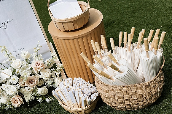 Wedding fan basket filled with paper fans and ceremony program cards beside a floral welcome sign on a grass lawn outdoors