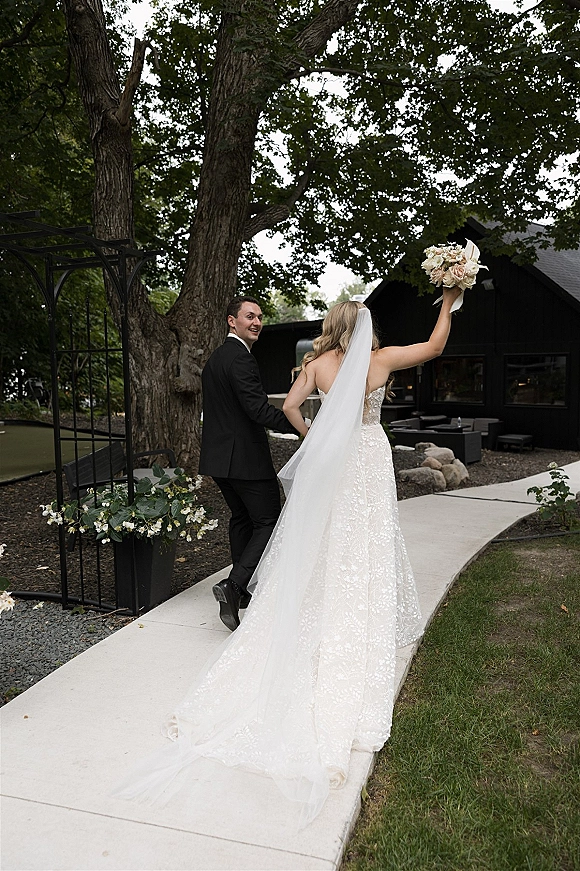 Wedding couple portrait of bride and groom walking away hand in hand, bride lifting a blush and white bouquet on a garden walkway by a modern black venue