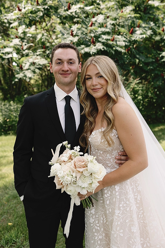 Couple portrait of bride and groom embracing outdoors, bride holding a white and blush bouquet with veil, trees and lawn behind
