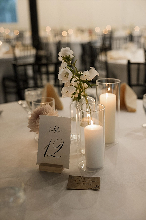 Wedding tablescape with wedding table number cards, white pillar candles and bud vases of white flowers and greenery on a white tablecloth indoors