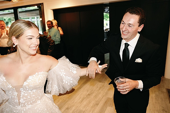 Wedding reception dance as bride in puff-sleeve gown laughs while leading groom in black suit holding cocktail glass on busy dance floor