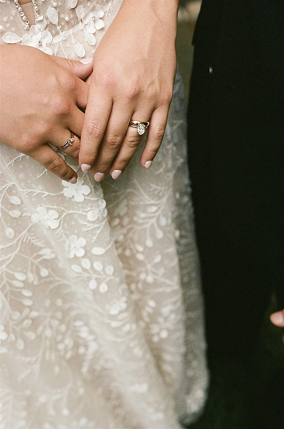 Wedding rings close-up on bride’s nude-manicured hand, showing an oval diamond engagement ring and band against lace dress detail