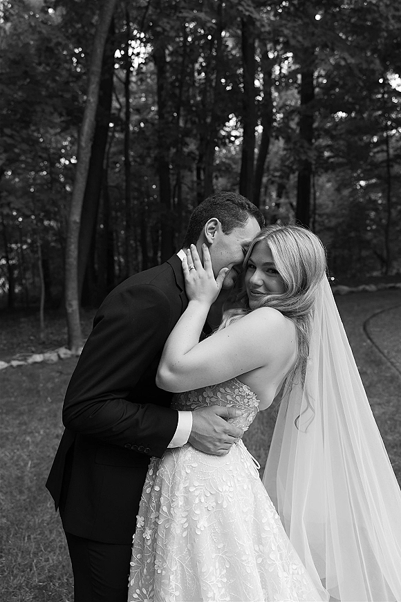 Couple portrait, black and white wedding portrait of groom whispering as bride looks at camera, long veil and tuxedo on a forest path