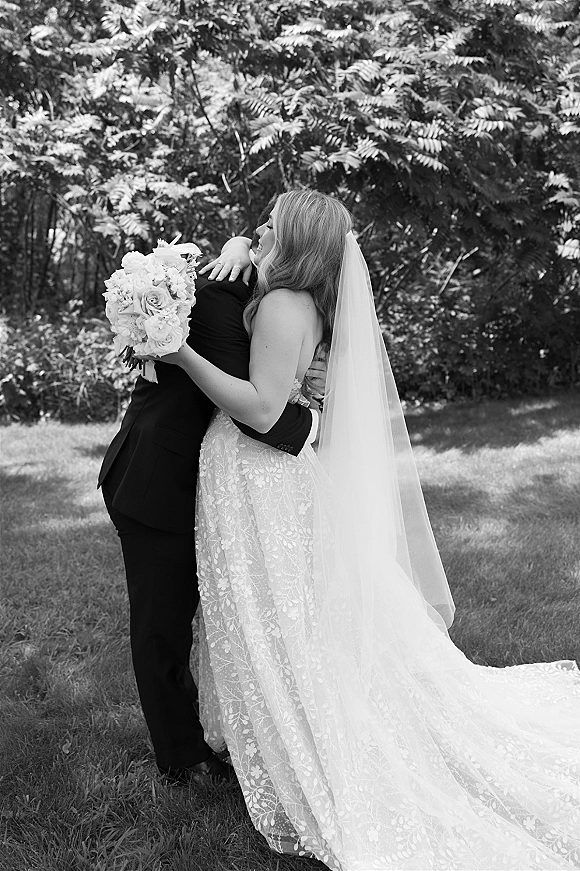 Wedding hug as bride and groom embrace on a grassy lawn with trees behind, bride in lace dress holding bouquet, groom in tuxedo