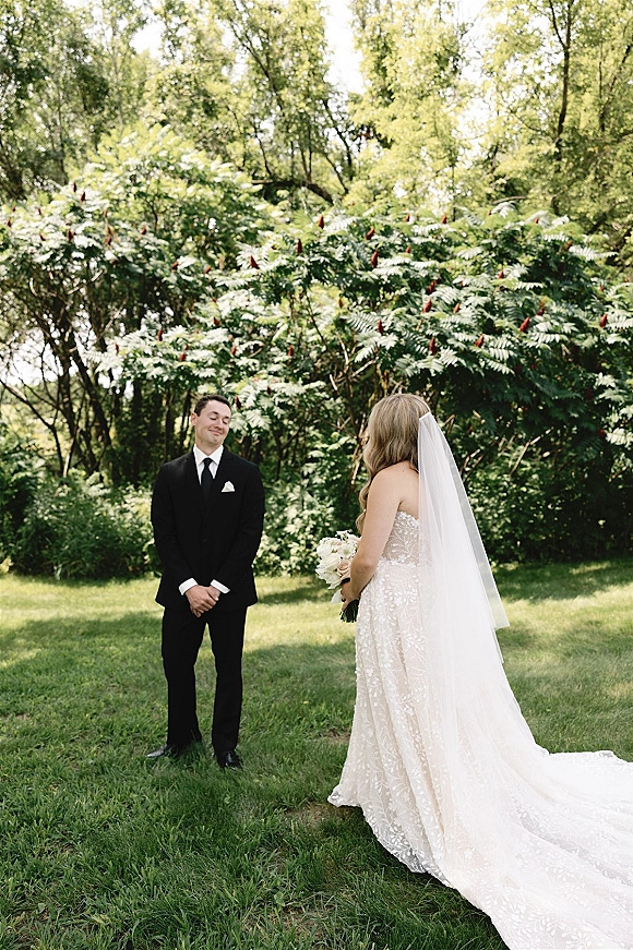 First look moment as bride in lace wedding dress and veil approaches groom in black tuxedo, holding bouquet on sunlit lawn among trees