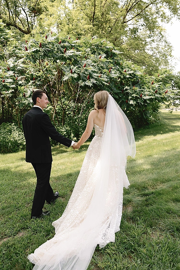 Couple portrait of bride and groom holding hands, walking on a tree-lined lawn in daylight, her long veil trailing behind his tuxedo
