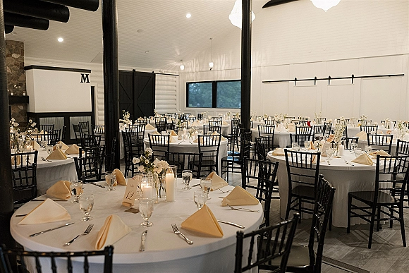 Reception tablescape with round wedding reception tables, white linens, black Chiavari chairs, beige napkins, and floating candle vases with florals in a modern barn hall