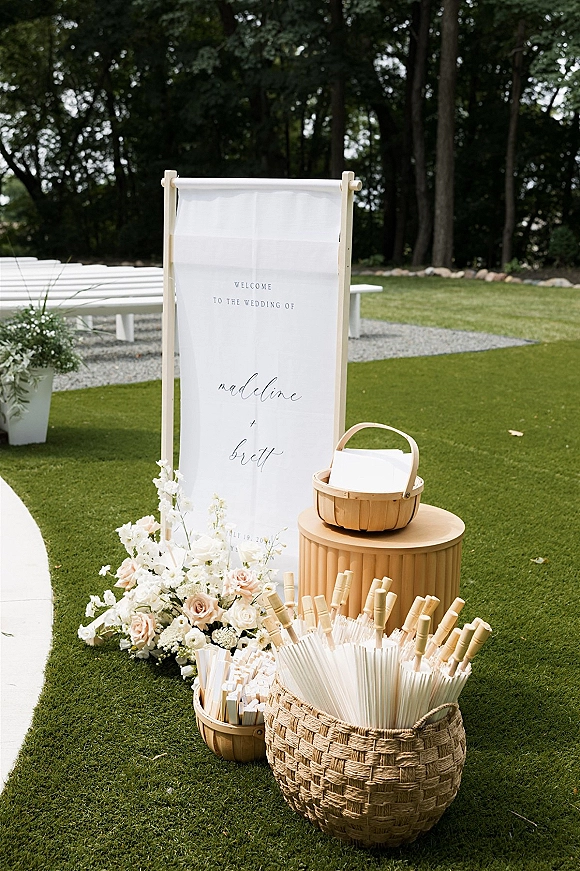 Wedding welcome sign on a wooden stand with a fabric wedding welcome sign and parasols in a wicker basket beside florals on a lawn aisle