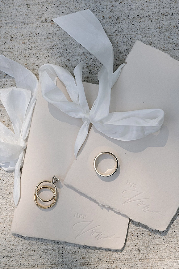 Wedding rings and engagement ring with band resting on handmade paper vow booklets tied with a white ribbon on a stone surface